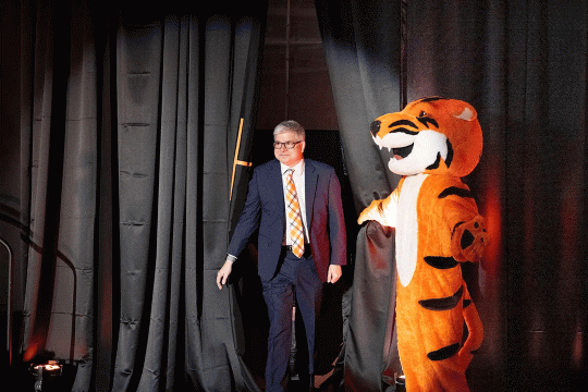 Mascot Ritchie pulls back a black stage curtain for William Sanders, RIT's next president, so he can walk onto the stage.