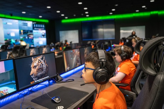 young students sit in a computer lab using a program and wearing headsets.