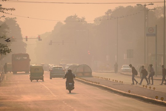 The photo shows a busy street scene. Three cars and one motorcycle are driving on the road, appearing on the left side of the image, and a group of four people walk across the road on the right side of the image. The air is heavy with brown/red color smog, and more people and cars can barely be made out in the distance. It is difficult to see much in the scene due to the air pollution.