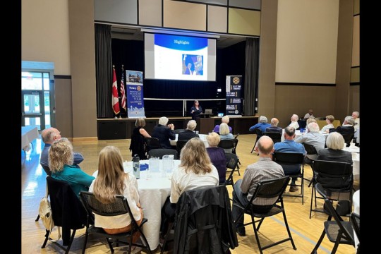 A room full of people seated at round tables, most of them seem to be older in age with white and gray hair. At the front of the room is a small stage and projector screen. In front of the stage, Sarah Burns stands in front of a podium with her laptop during her presentation.