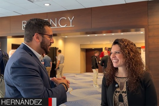 A screenshot of the news interview. The male reporter stands on the left in a blue suit, and Rachel Conidaris stands on the right in a black jacket and brown shirt. Both as smiling as they converse.