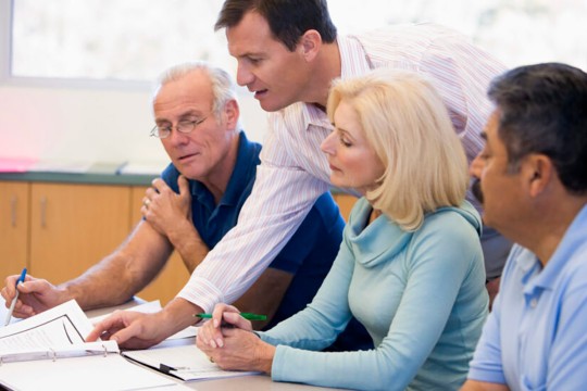 Older adult learners sit at a desk together with a professor leaning over them showing them something in a notebook.