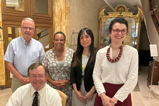A group of people stand behind Gerry Buckley as he signs paperwork on a desk. They are all smiling. 