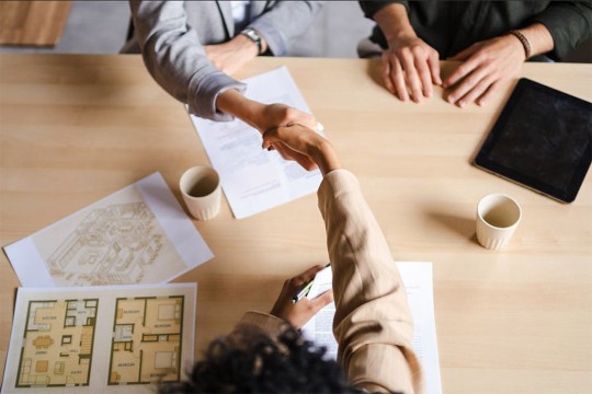two people shake hands over a table with paperwork spead throughout the frame.