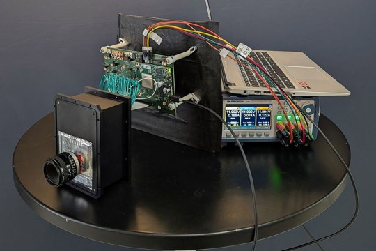 A complex set of electronics on a table, including a sensor in a black box, a laptop, and interconnected cables.