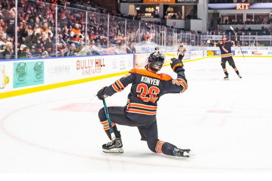 A hockey player takes a knee on the ice and faces the crowd, giving the cheering group a wave. There is another RIT hockey player in the background.