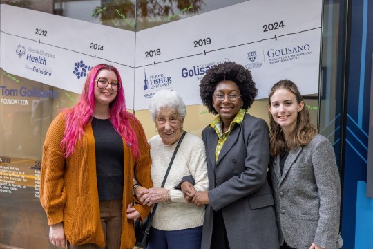 4 people stand in front of a banner bearing the Golisano name.