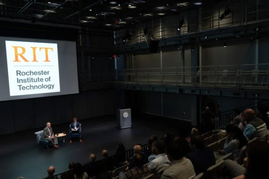two men sit on a stage with the R I T Logo in the background.