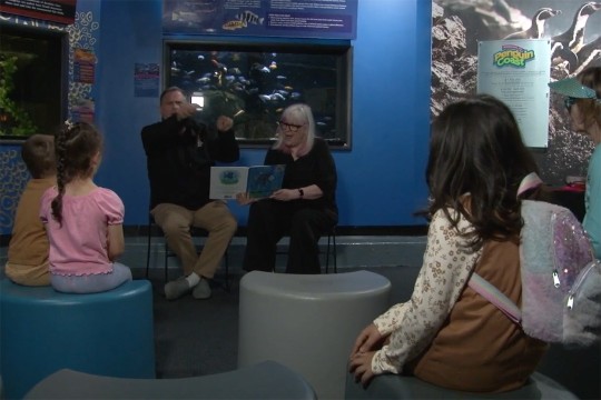 a man signs to children sitting in a large room at an aquarium.