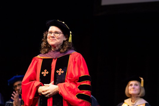 a woman in glasses and curly brown hair stands in red regalia for the university on a stage.