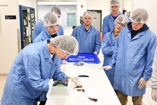 One high school student splits a silicon wafer at a table while six other high school students observe in a clean room facility.