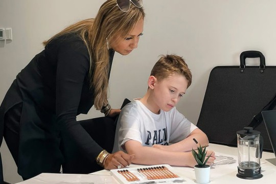 a young boy sits at a table illustrating a picture while his mother looks over his shoulder.