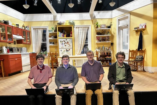 'four students sit on a stage with the set for a play behind them'