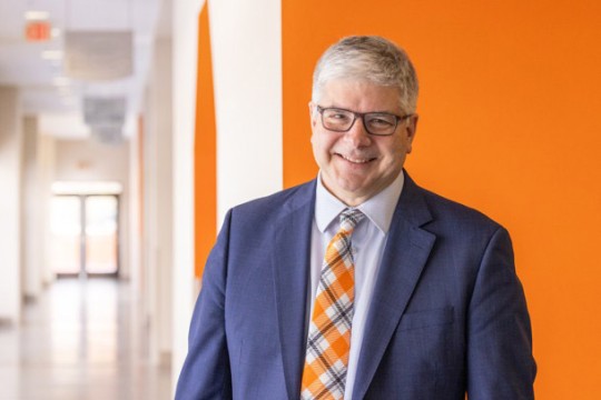 R I T President Bill Sanders stands in a suit and tie in front of an orange background in a brightly lit hallway.