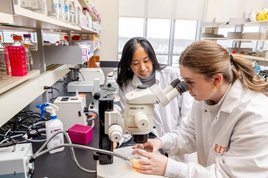'two women stand next to each other in a research lab with one of them using a microscope.'