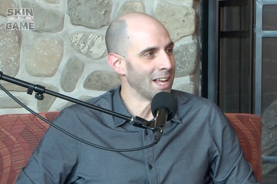 'a man in a gray button down shirt sits in a red chair speaking in to a microphone in front of him.'