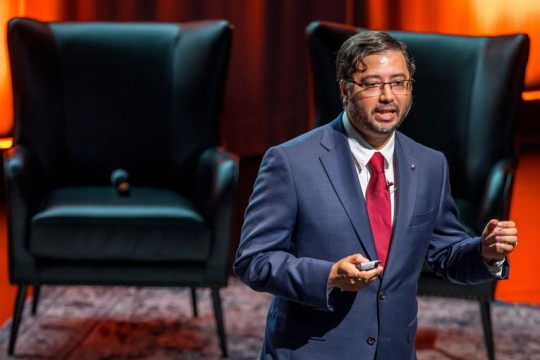 a man in a blue suit and red tie stands on a stage speaking to a crowd during a Ted X talk.