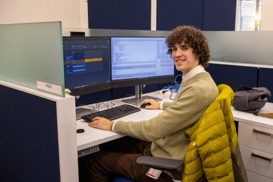 'a student in a tan sweater sits in a cubicle in front of a computer screen'