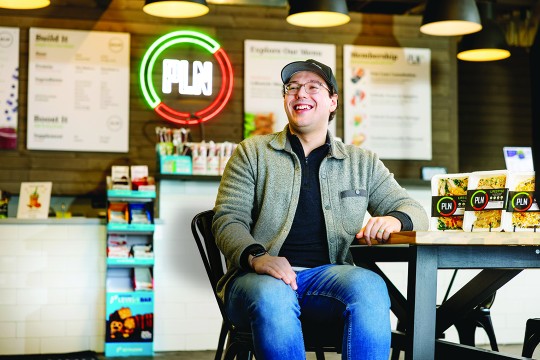 'a man in a gray zip up shirt and baseball cap sits at a table in a casual industrial restaurant space.'