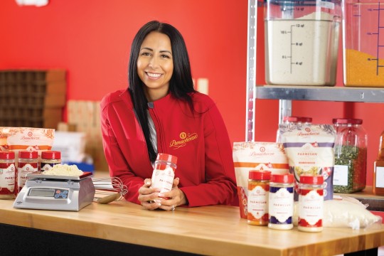'a woman in red stands against a butcher block countertop in an industrial kitchen holding a bottle of shredded cheese product.'