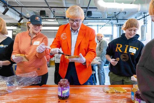 RIT President Bill Sanders and his wife, Emily, spread jelly on a piece of bread. A student stands next to them spreading peanut butter on the pre-jellied bread. All of them wear RIT orange hairnets.