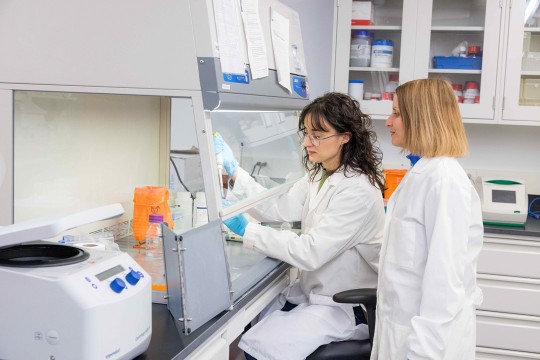 'two women stand next to each other in a research lab with one of them holding a pipette.'