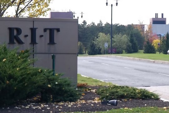 a cement fence with the R I T logo is shown near an entrance to the campus.