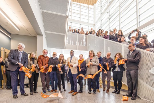 a group of people stand in a modern building cutting a large orange R I T ribbon with large scissors.