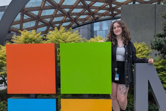 'a college age woman stands next to a multi colored cube on the Microsoft campus.'