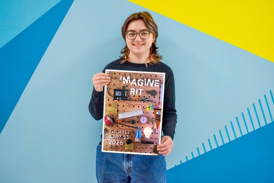 'a person with brown wavy hair and glasses in a black shirt and jeans stands in front of a colorful wall holding an Imagine R I T poster they designed.'