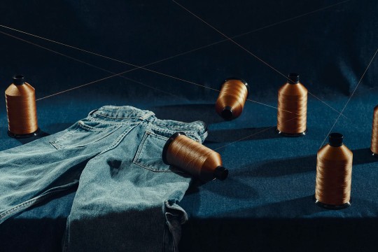 a pair of jeans and several spools of gold thread sit on a blue table against a dark background.