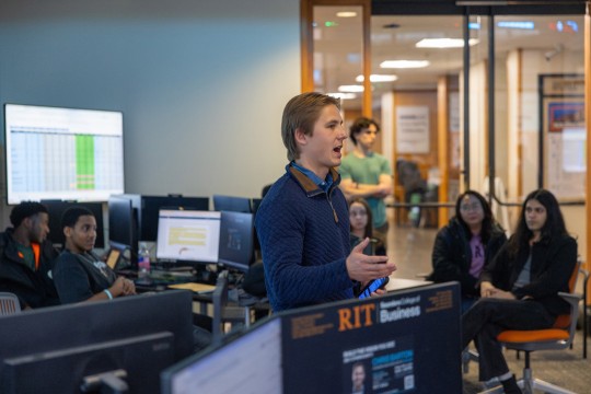 a young man stands in a finance lab speaking to a club