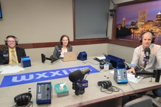 three people sit at a table that says W X X I. They are all wearing headphones and sit in front of microphones.