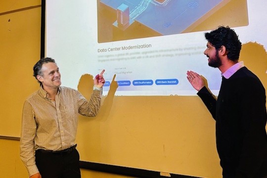 Prof Martinez in checkered button up gestures toward projector screen where "Data Center Modernization" case study is visible. Another student stands to his right also gesturing.