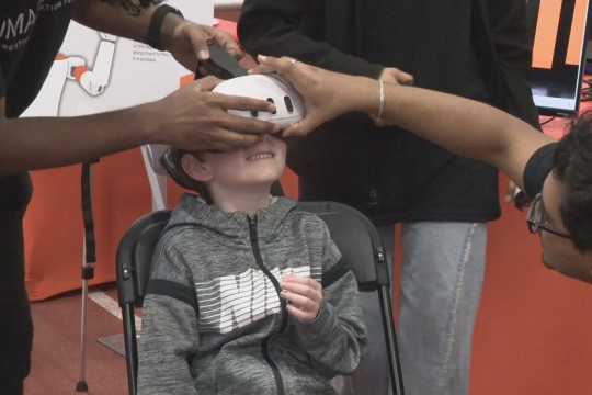 a young boy sits in a chair while two adults fit a V R helmet to his head.