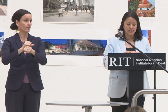 two woman stand at the head of a room, one interpreting and one speaking in front of an R I T N T I D podium sign.