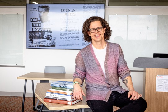 'a man with a pink cardigan and shoulder length curly hair wears glasses and leans on a stack of books placed on a desk in a classroom.'