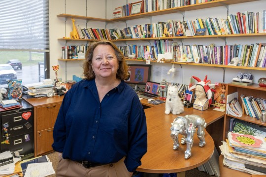 a woman with curly brown hair and a blue button down shirts leans against a desk in a small office filled with books and knick knacks.