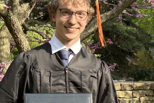 Student posing in front of flowering tree holding diploma, dressed in cap and gown