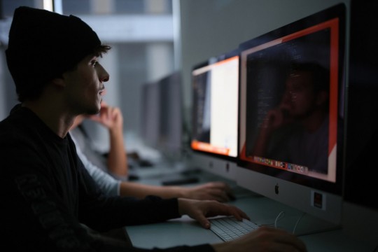 a man in a black hat sits in front of a computer in a darkened room