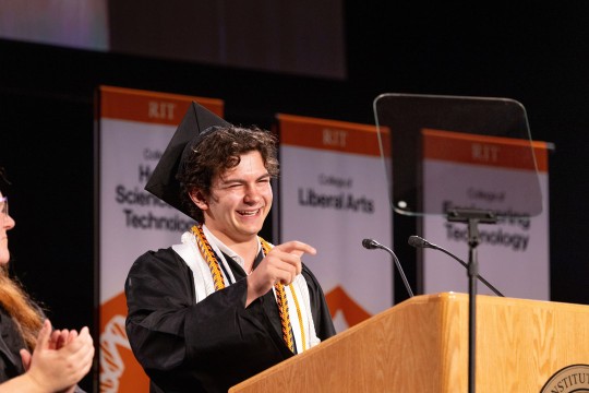 A student laughs while standing at a podium in commencement regalia.