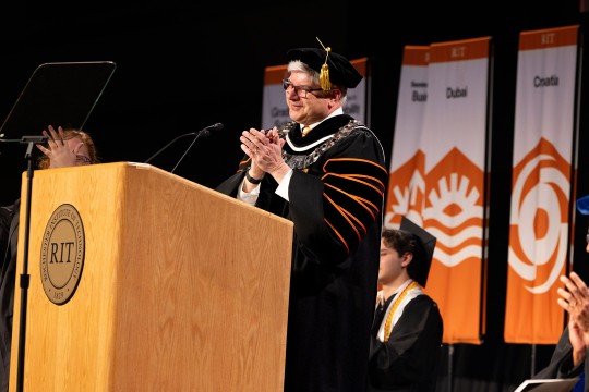 University president claps his hands while standing at podium in full academic regalia