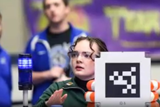 A woman in safety glasses sits next to a blue flashing light and QR code.