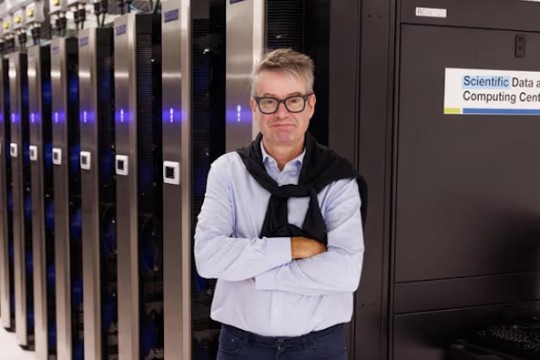 a man in a blue shirt with a navy blue sweater over his shoulders stands in front of a server farm