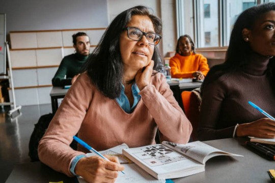 an older woman in glasses sits at a desk in a modern classroom.