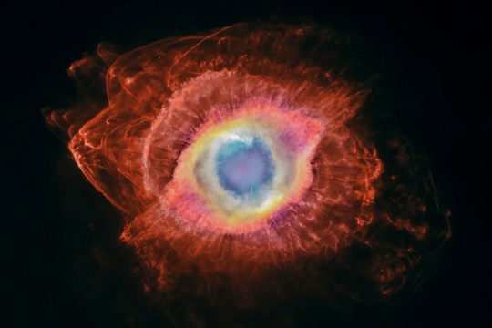 A telescope image of a colorful nebula with red material seeming to emanate from a blue, yellow and pink eye at the center.