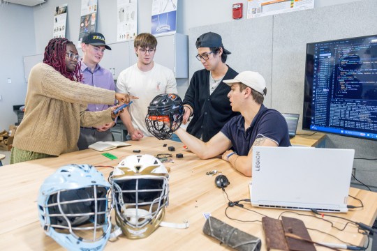 'five students sit in a workshop, working collaboratively on a specialized lacrosse helmet.'
