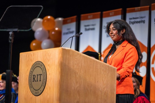 'a student orange blazer as she welcomes the audience at the Gordon Field House and Activities Center for New Student Convocation'