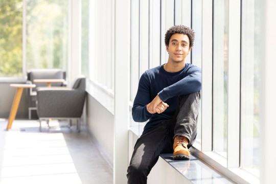 'a college age male sits against a window in a brightly lit atrium with casual seating in the background.'