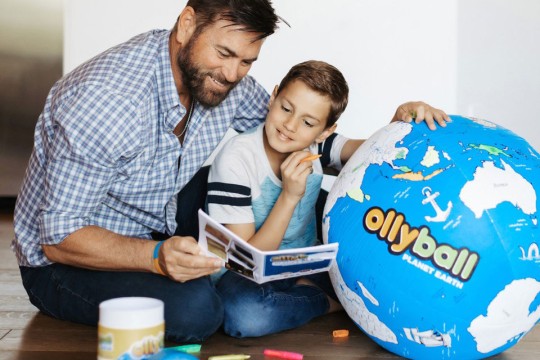 'a man and a young boy sit on the floor next to a large ball designed to look like a world map that reads Ollyball on the side.'